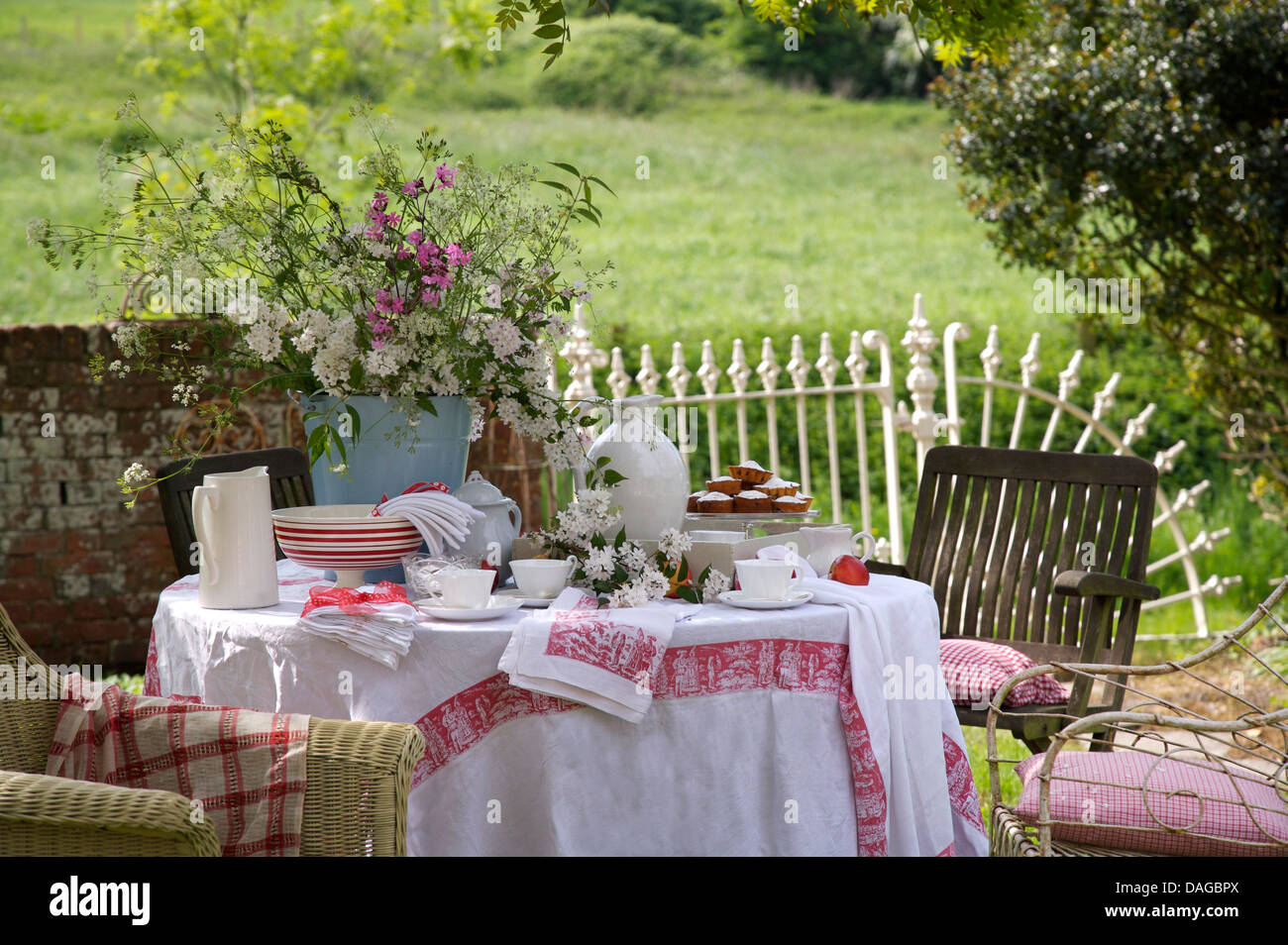 Lawn in country garden with vase of summer flowers on table with jug and bowls and  white linen cloth edged with red Stock Photo