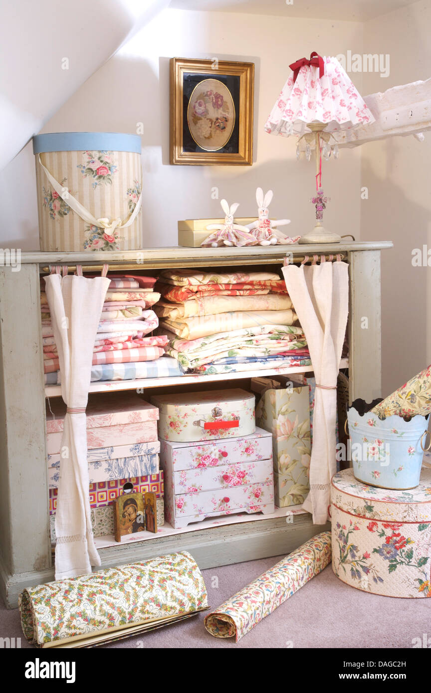 Corner of cottage bedroom with folded linen and floral boxes on shelves in open cupboard with white cotton drapes Stock Photo