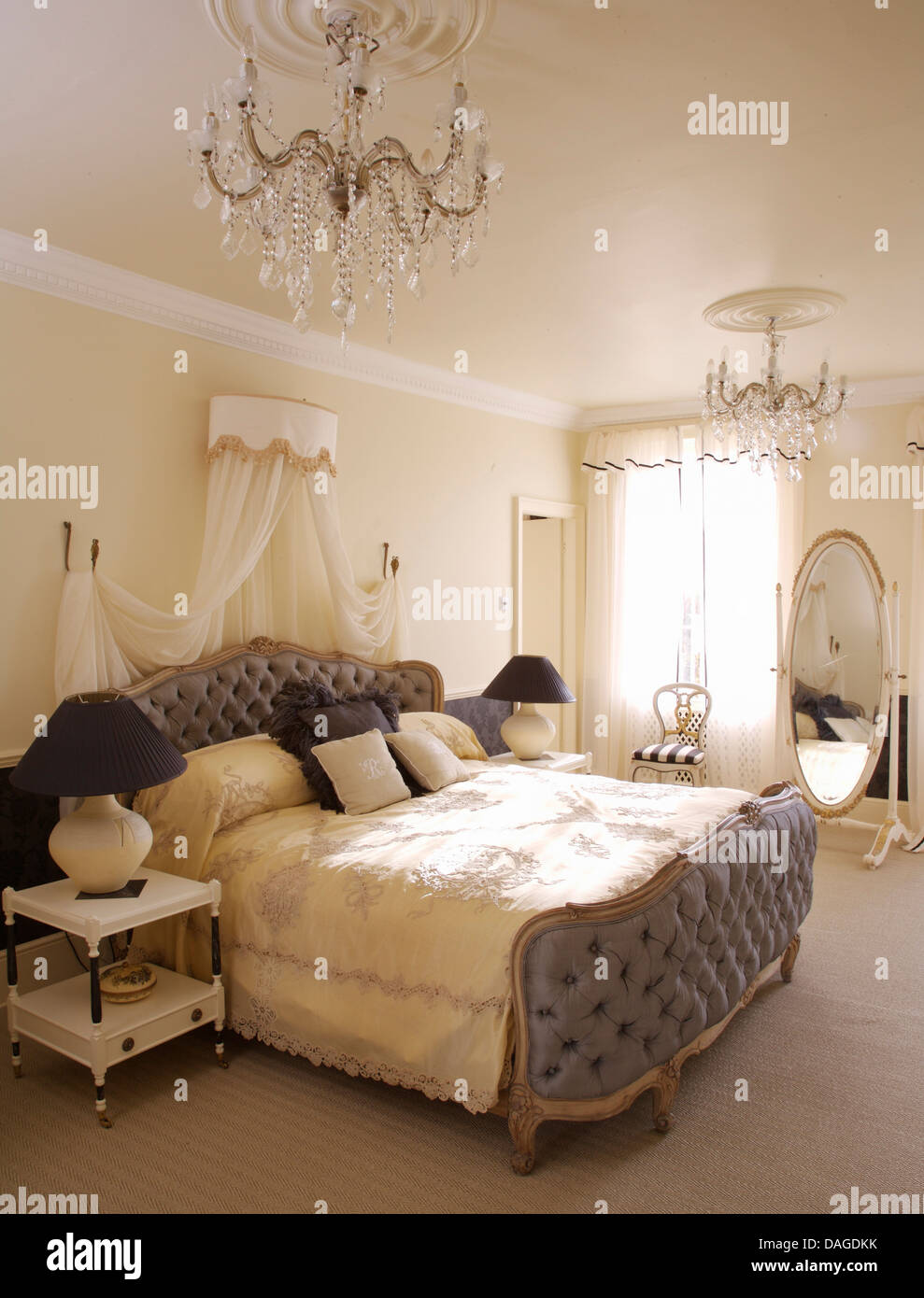 Upholstered double bed with white voile drapes in opulent bedroom with glass chandelier and cheval mirror Stock Photo