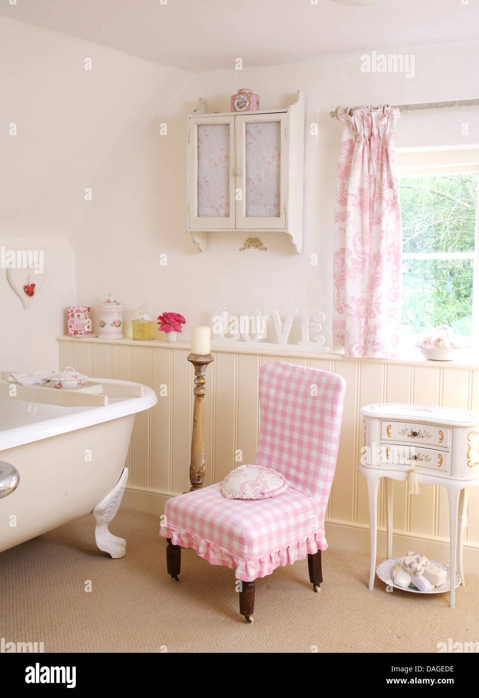 Pink checked loose cover on chair next to roll top bath in country bathroom with white wall cabinet and pink curtains Stock Photo