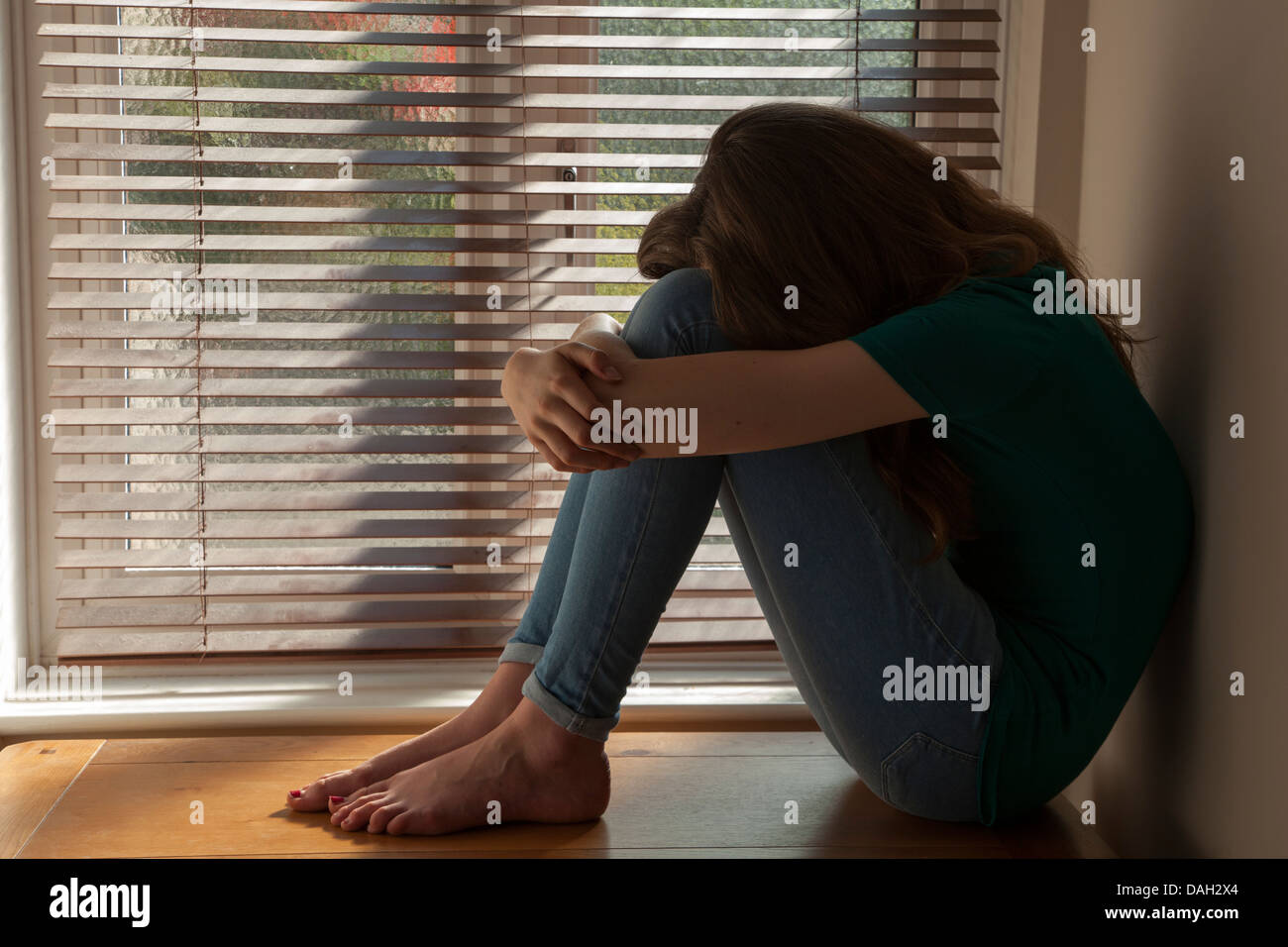 A teenage girl, head between her knees, sitting by a window with light