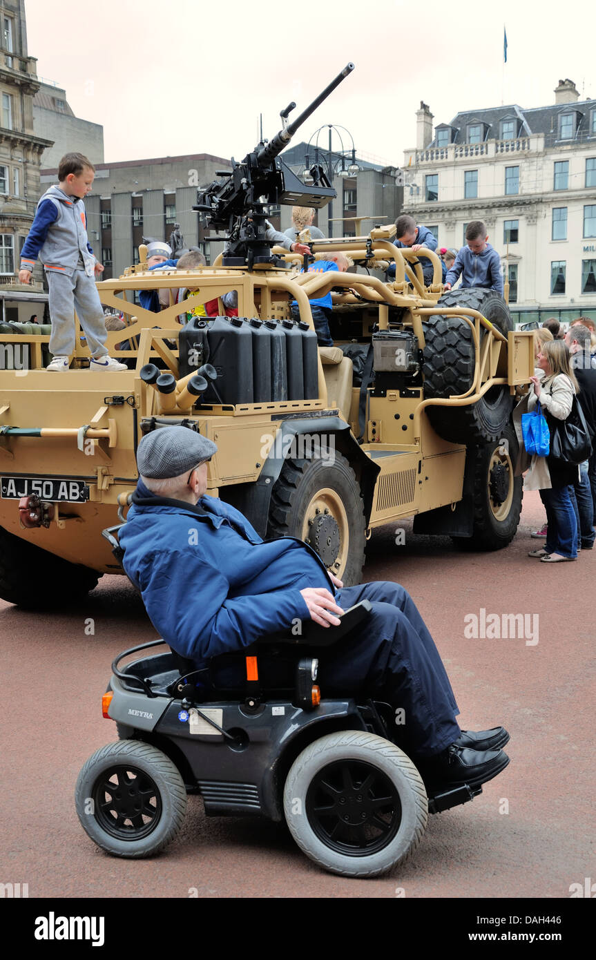 Man in mobility chair stops to watch children playing on an armoured car on armed forces day in Glasgow, Scotland, UK Stock Photo