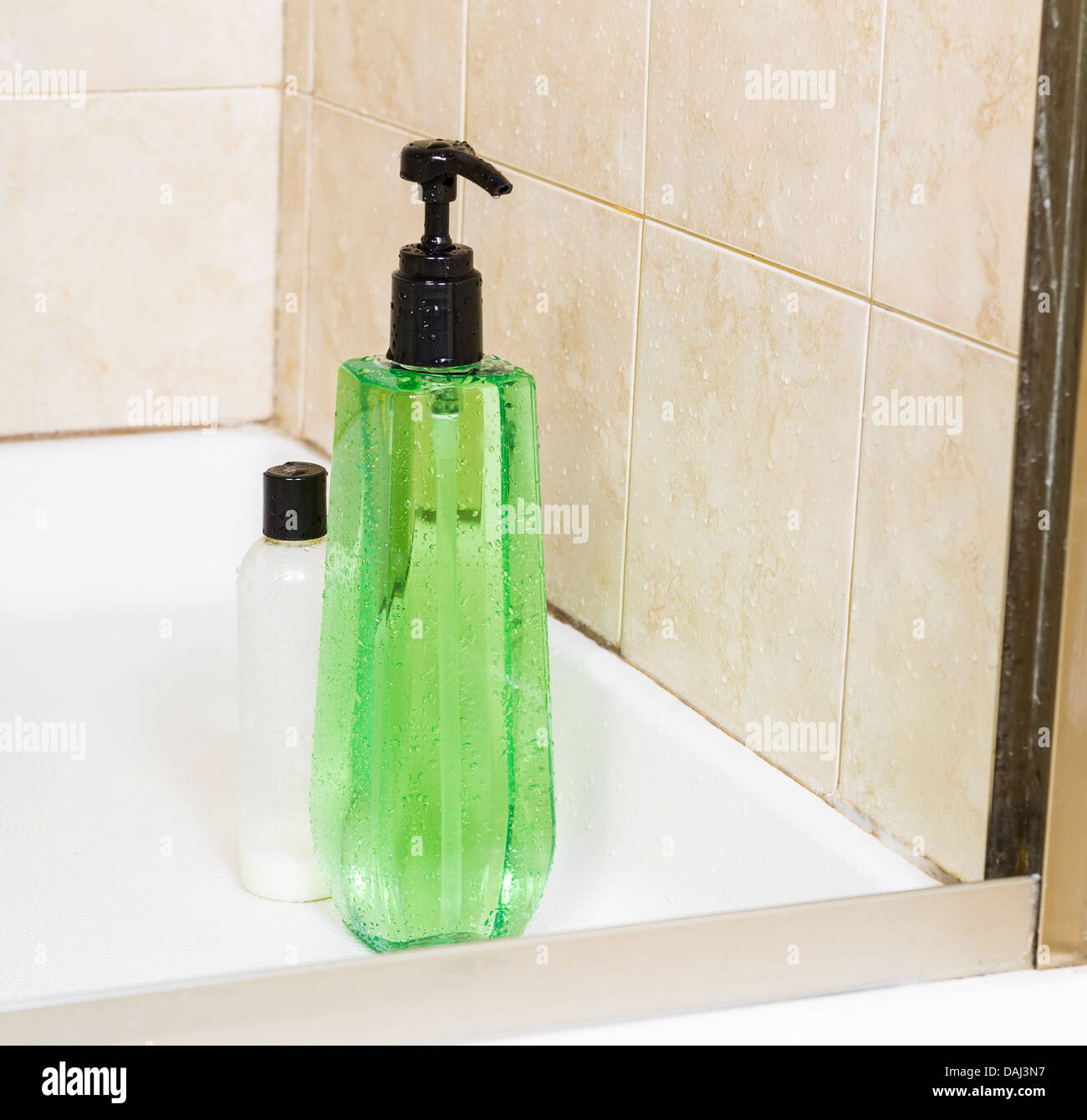 Photo of tall green shower gel bottle, with water drops, and a small bottle of hair conditioner with shower stall in background Stock Photo