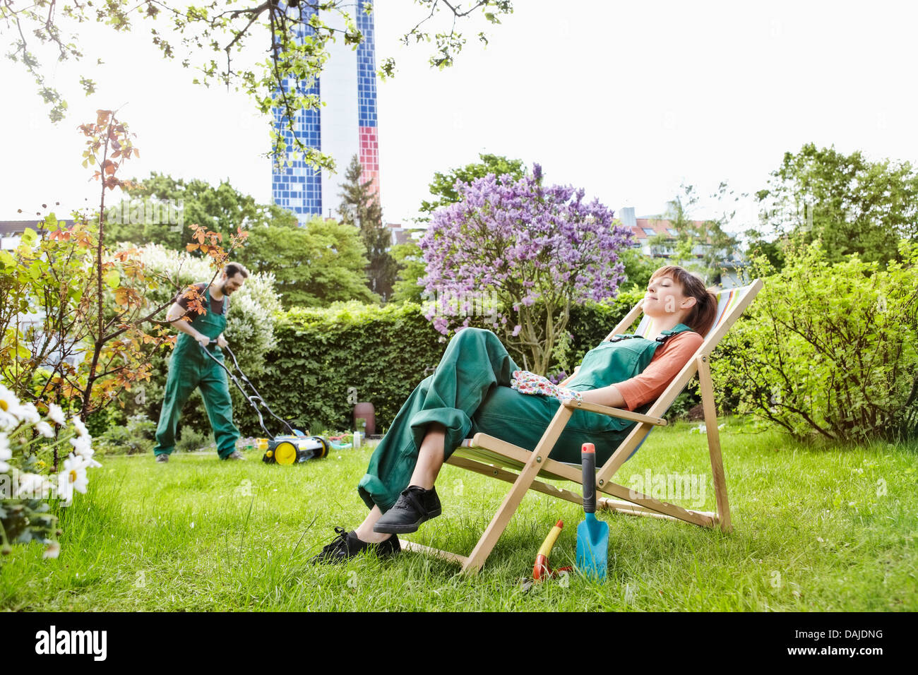 Germany, Cologne, Young woman relaxing on deck chair while man mowing lawn Stock Photo