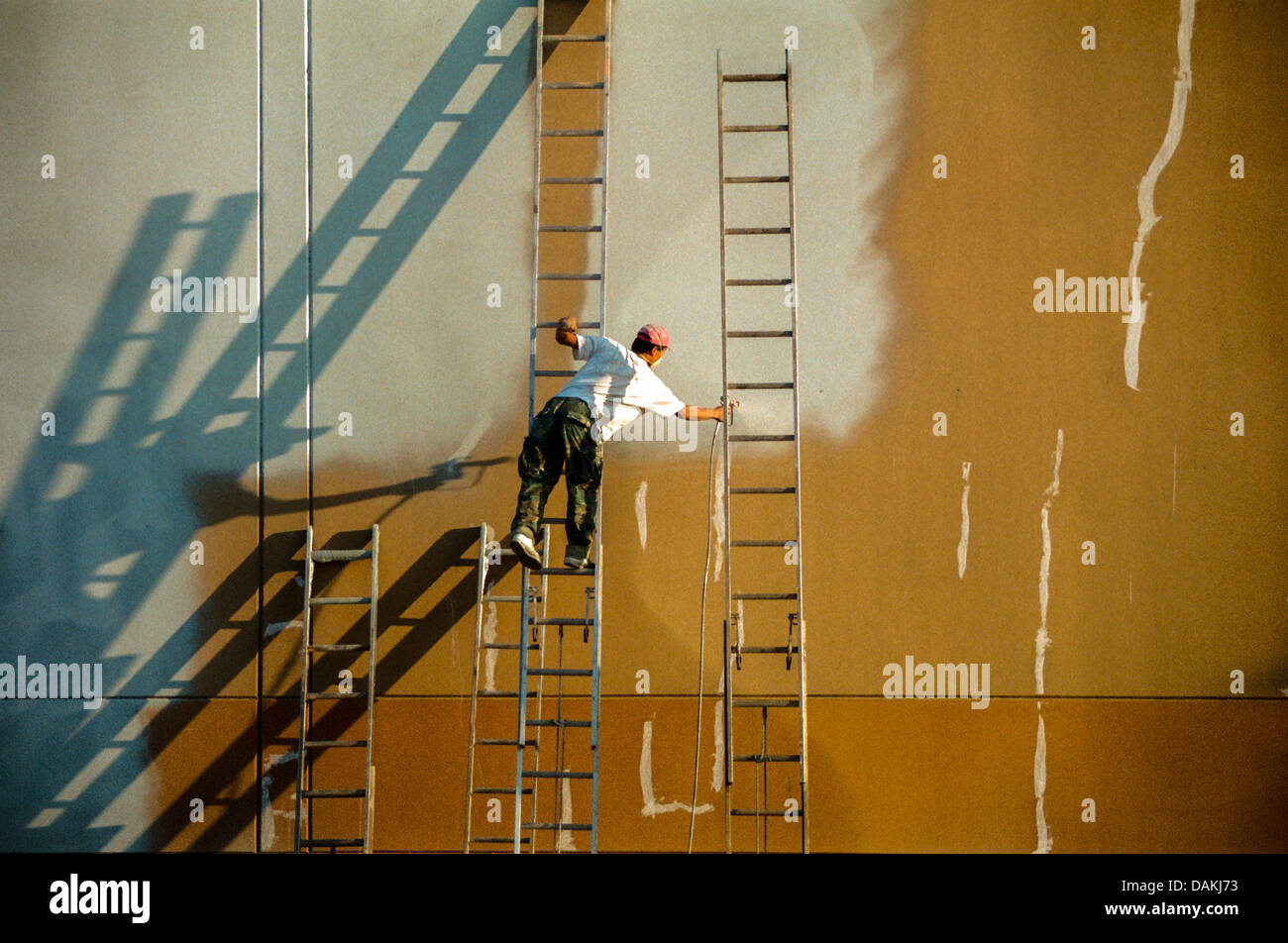 Balancing on a ladder, a Hispanic workman spray paints a factory wall in Irvine, CA. Note long shadows from afternoon sun. Stock Photo