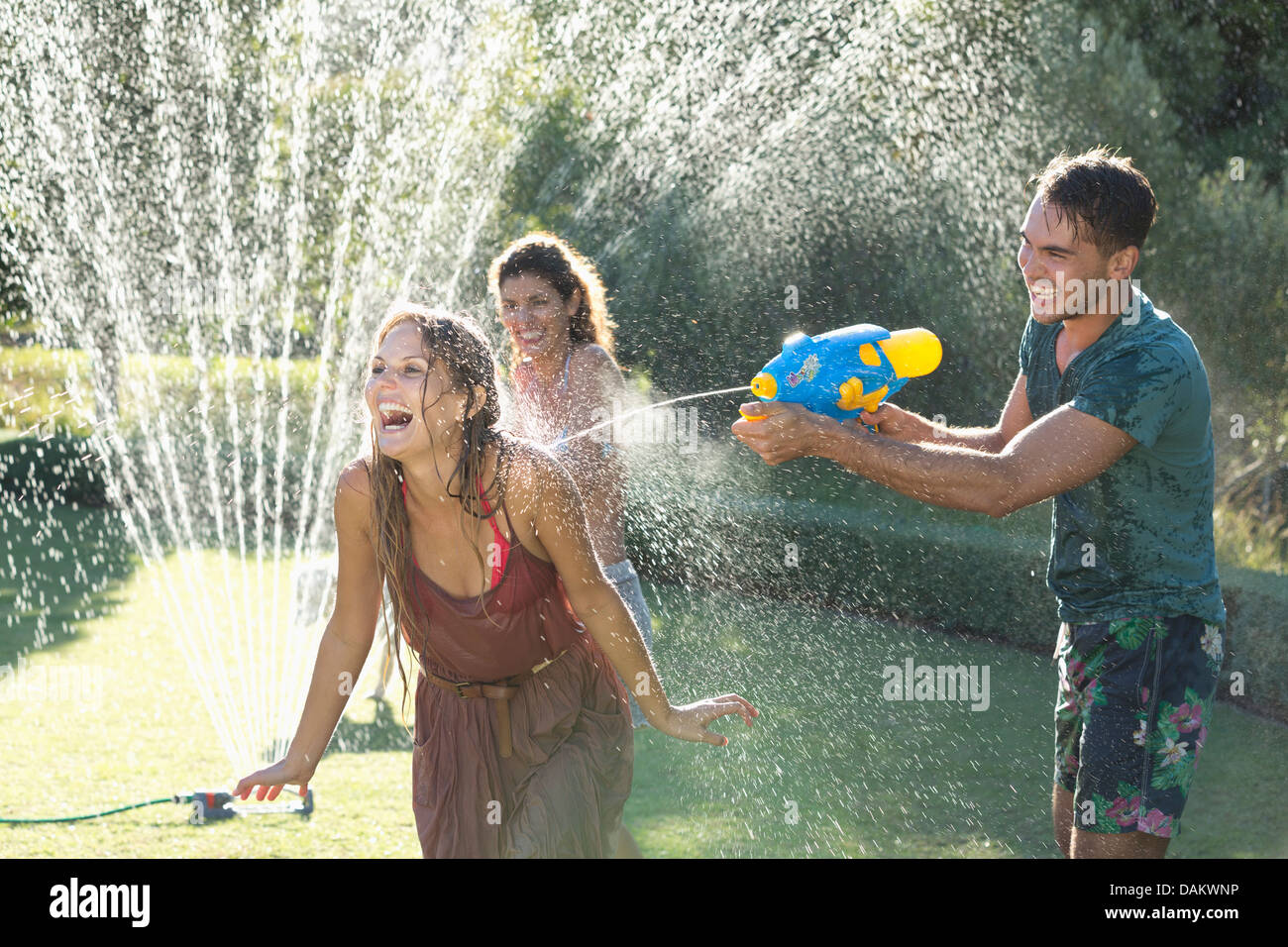 Friends playing with water guns in sprinkler in backyard Stock Photo
