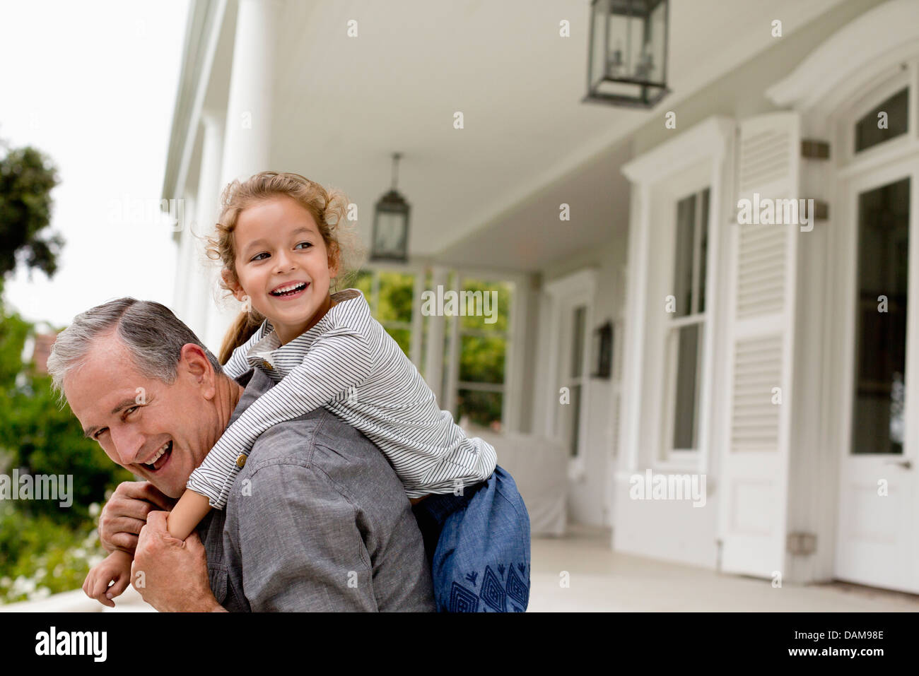 Older man carrying granddaughter piggy back on porch Stock Photo