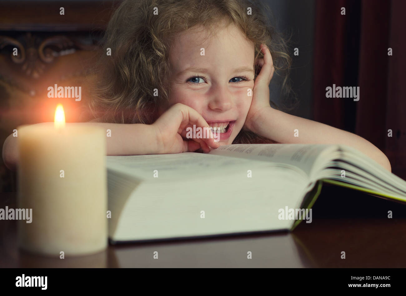 Cute little girl sitting in the dark with burning candle and reading a ...