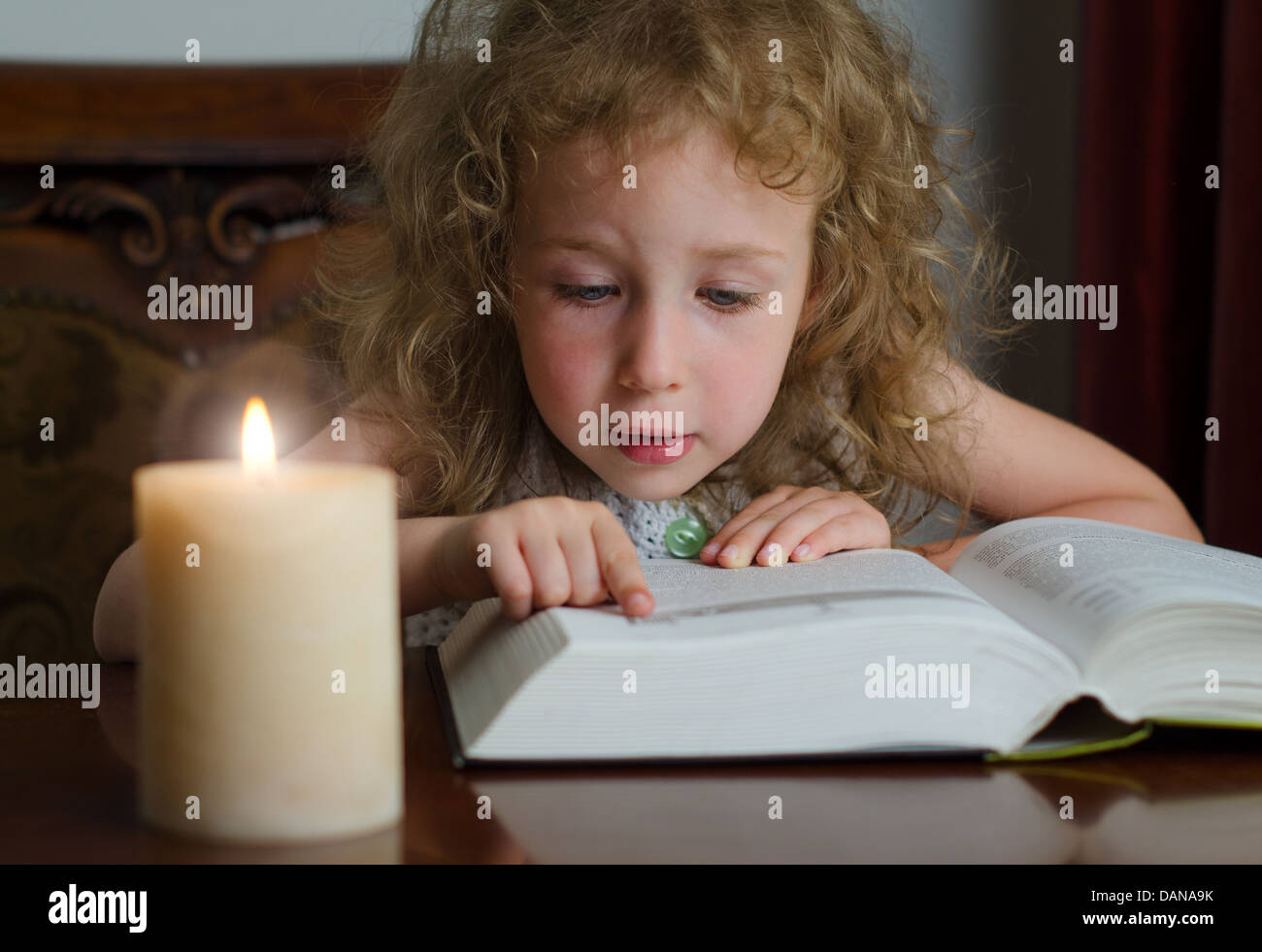 Cute little girl sitting in the dark with burning candle and reading a ...