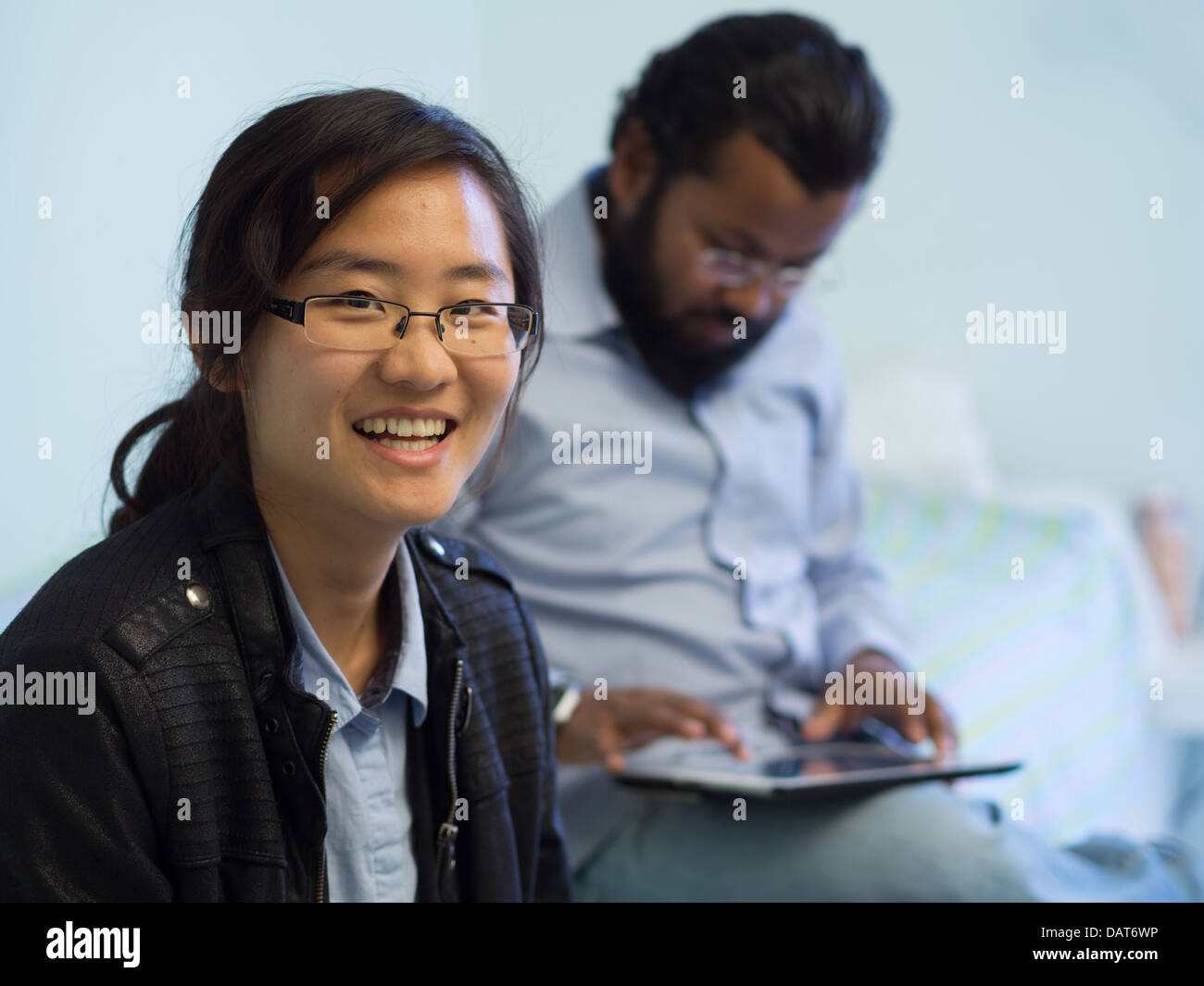 Portrait of an Asian woman smiling in foreground as an Indian man uses a tablet in the background Stock Photo