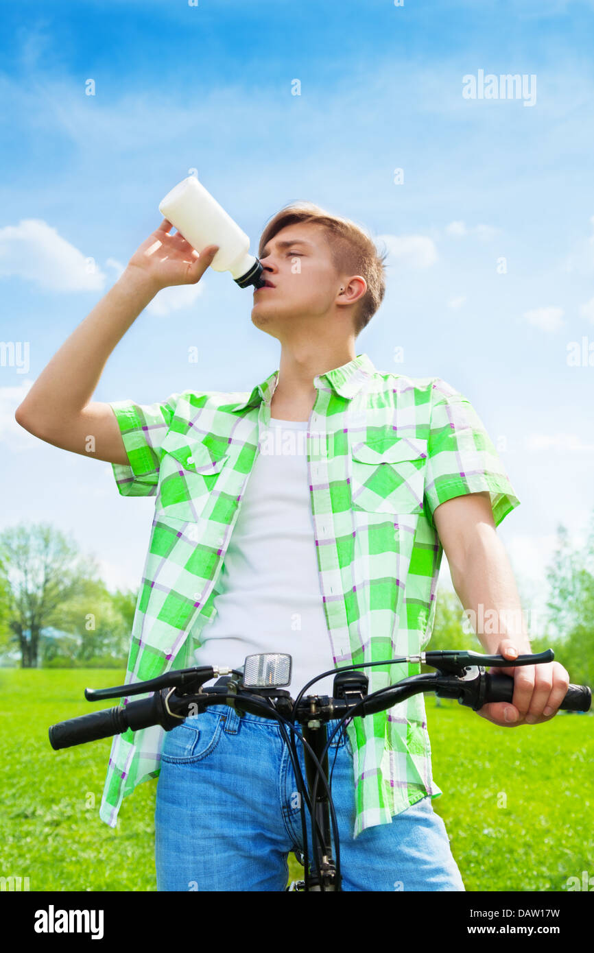 Young man sitting on a bike and drinking and fulfilling thirst with water in a bottle in a park Stock Photo
