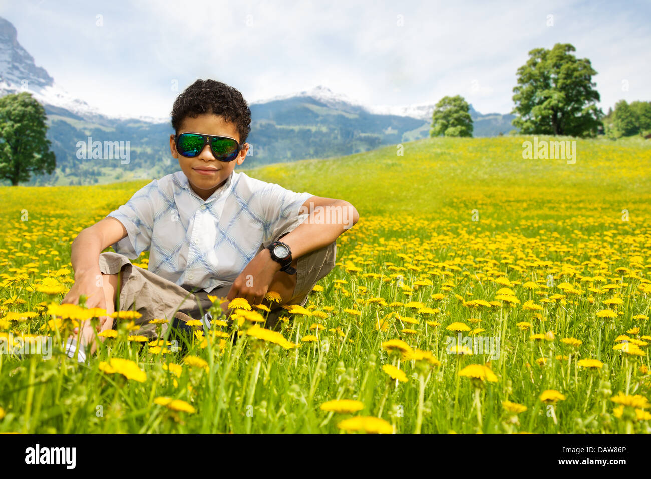 Happy little black boy in sunglasses sitting in the dandelion field with mountains on background Stock Photo