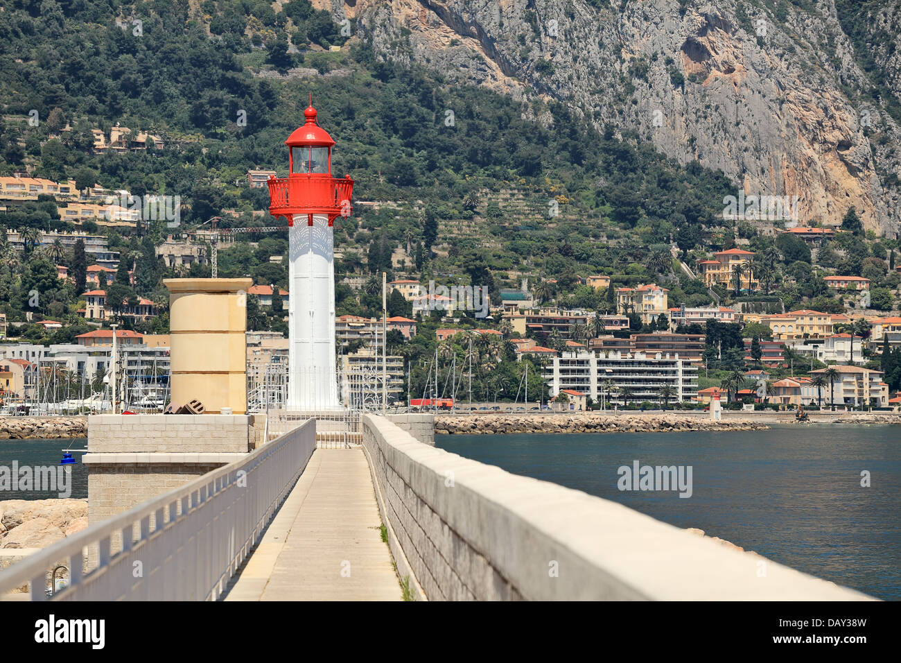 White and red lighthouse at the end of narrow promenade along the sea as houses and hotels on the slope in Menton, France. Stock Photo