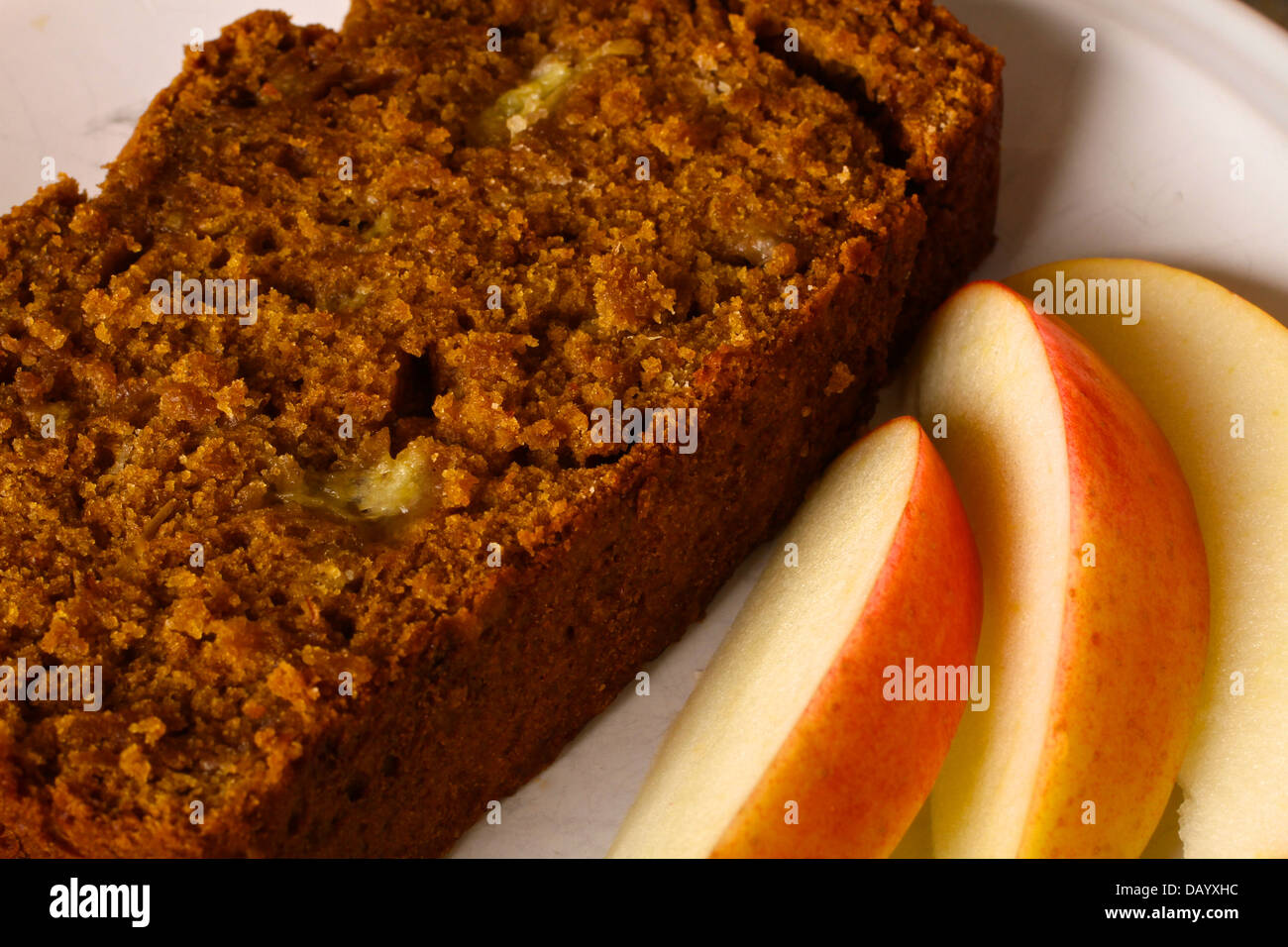 A slice of homemade banana nut bread is plated in front of a partially sliced loaf of bread, along with fresh apple slices. Stock Photo