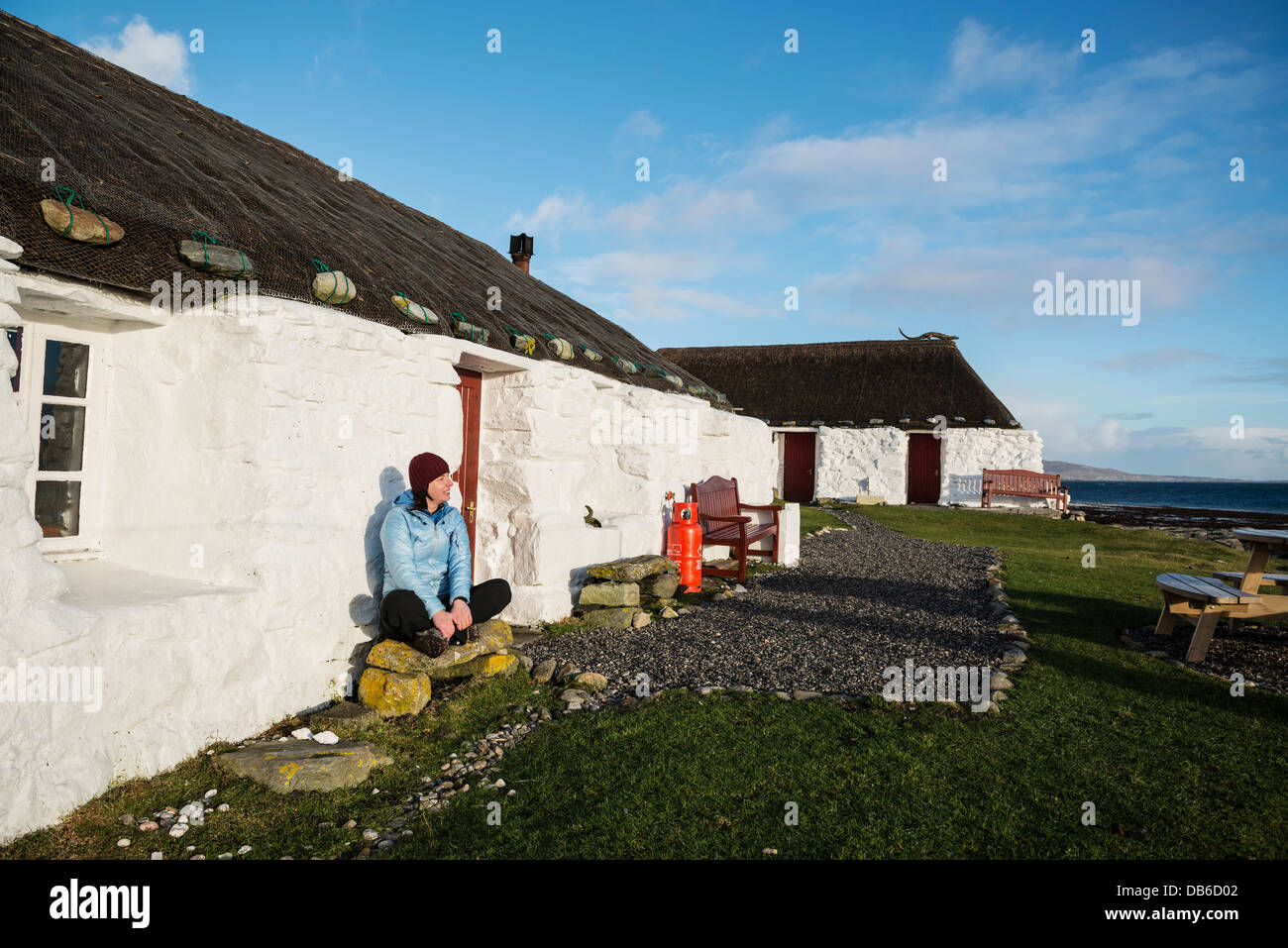 Worman sits infront of thatched roof traditional blackhouse now used as youth hostel, Berneray, Outer Hebrides, Scotland Stock Photo