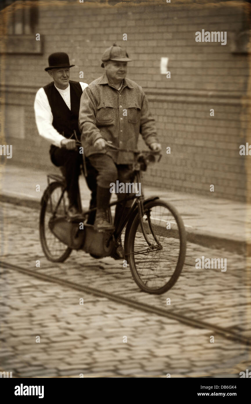 two edwardian men riding a tandem bicycle on a cobbled road Stock Photo