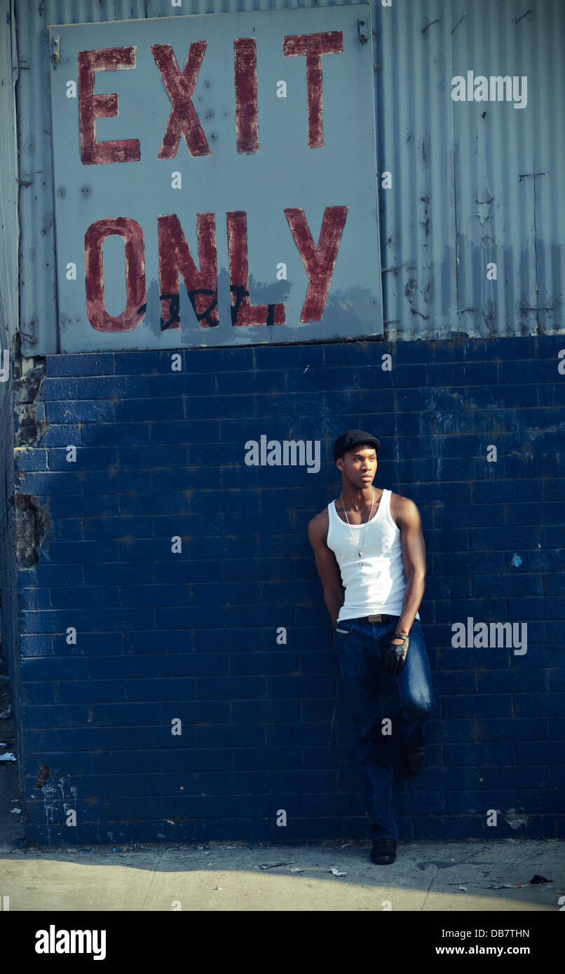a young attractive black man wearing a wife beater blue jeans leaning against a blue brick wall with exit only graffiti on wall Stock Photo