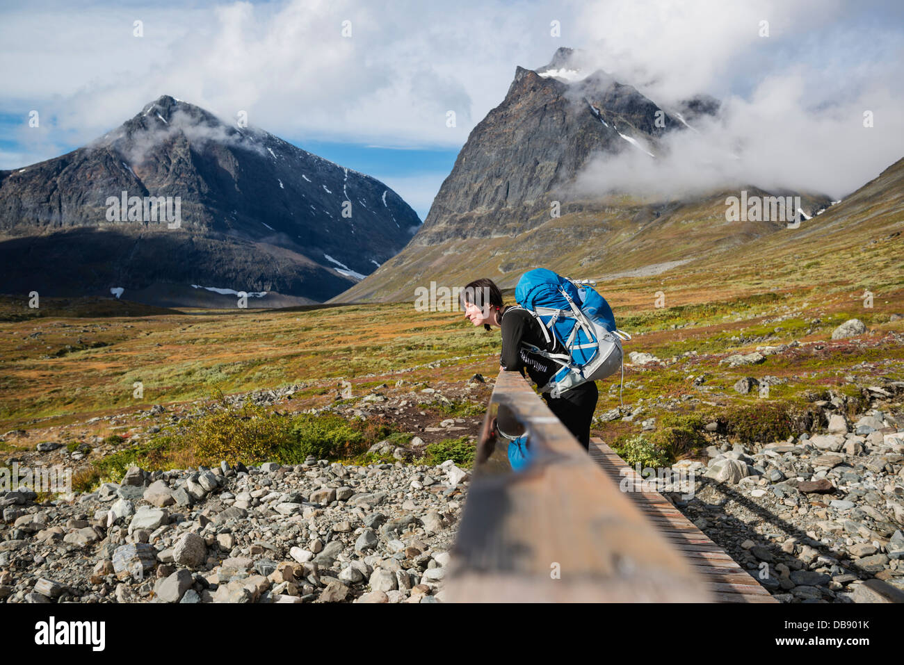 Female hiker leans on bridge in Ladtjovagge with Tolpagorni - Duolbagorni mountain in distance, Lappland, Sweden Stock Photo