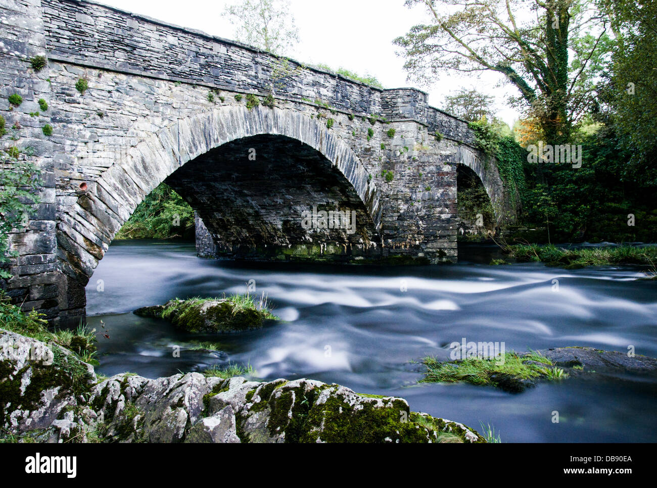 A long exposure shot of a stone bridge, with smooth river water running under and few rocks in the foreground. Skelwith bridge. Stock Photo