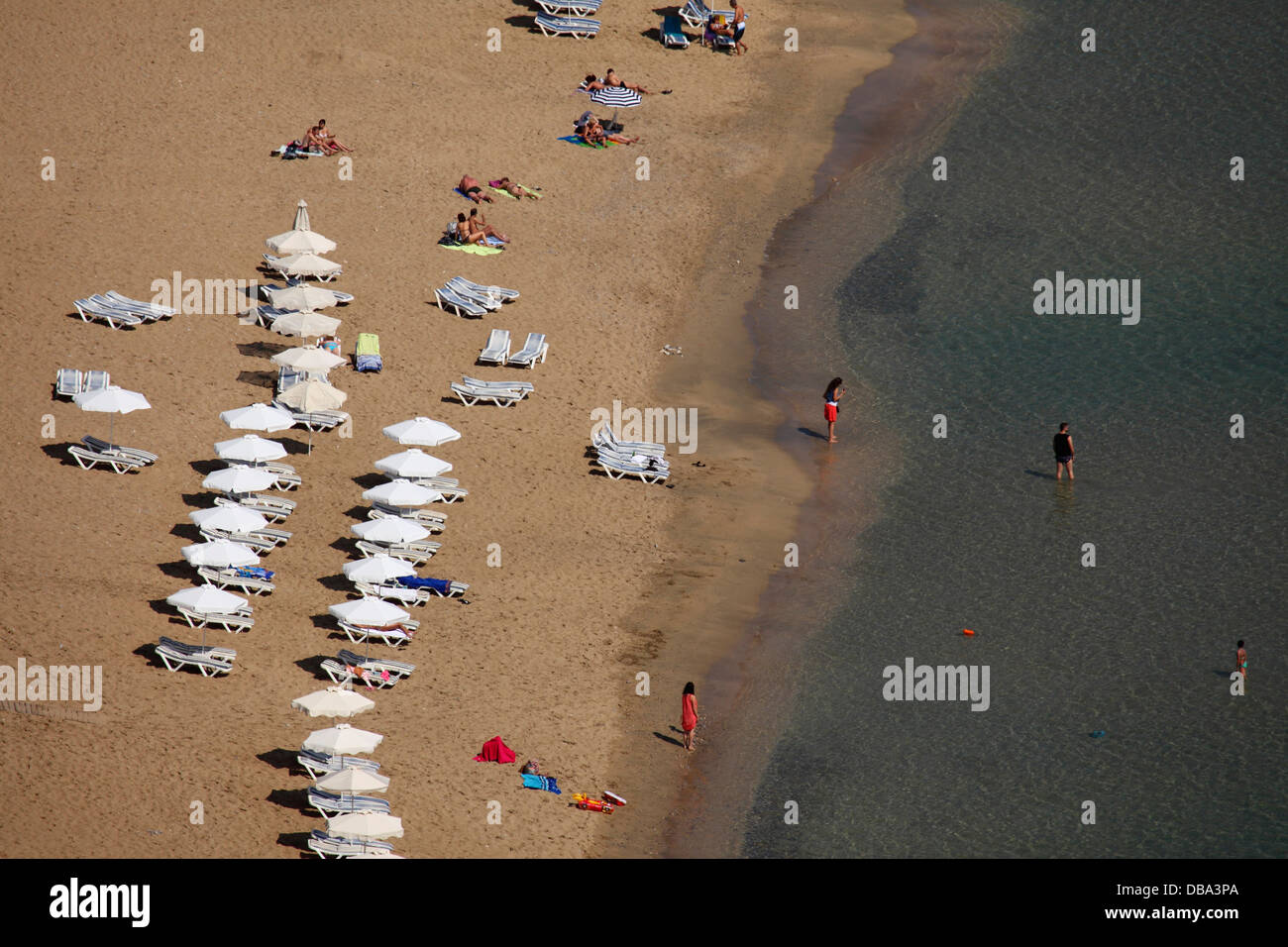 A beach holiday by the Agean Sea at Lindos, Rhodes, Greece. Sunbeds are lined up on the golden sand. Stock Photo