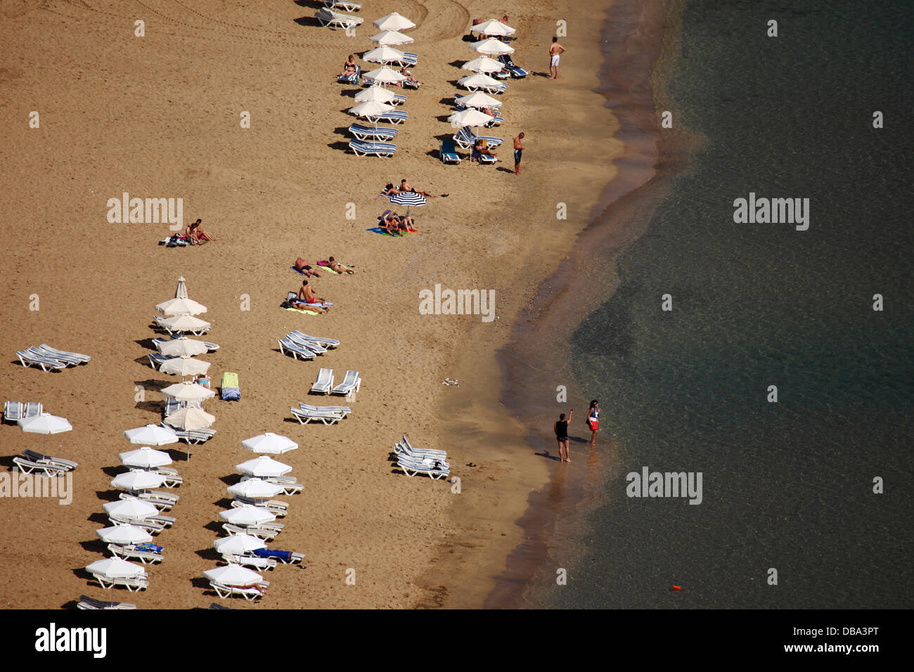 A beach holiday by the Agean Sea at Lindos, Rhodes, Greece. Sunbeds are lined up on the golden sand. Stock Photo