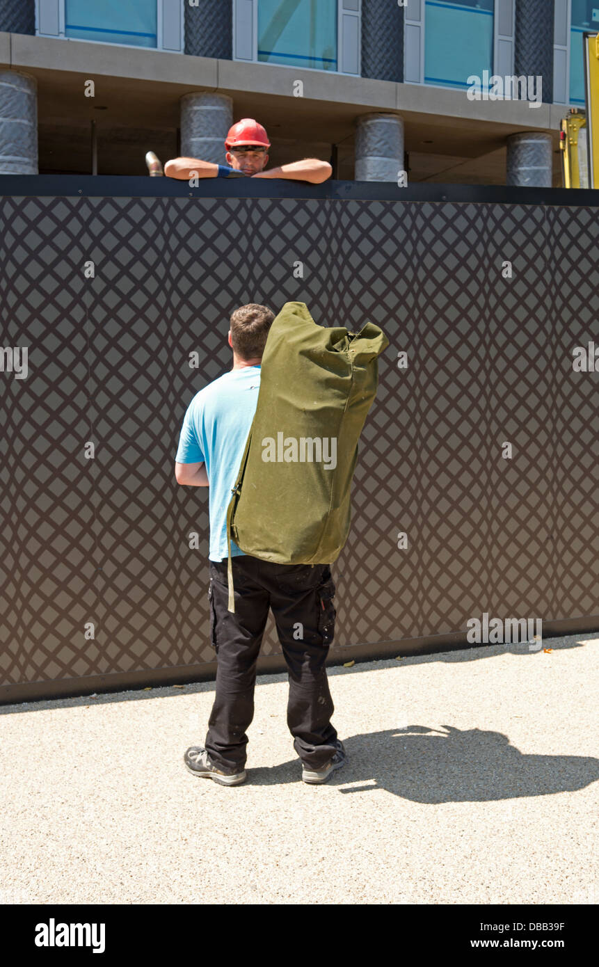 Builder talking over a fence to a man with a large kit bag. Stock Photo