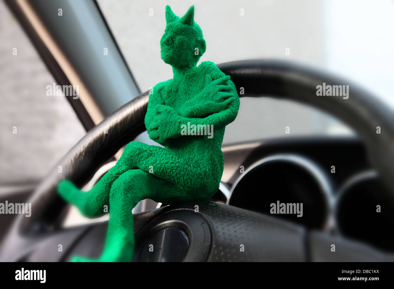 a little green devil made of clay, sits on the steering wheel of a car...he is waiting for someone to grab the wheel. Stock Photo