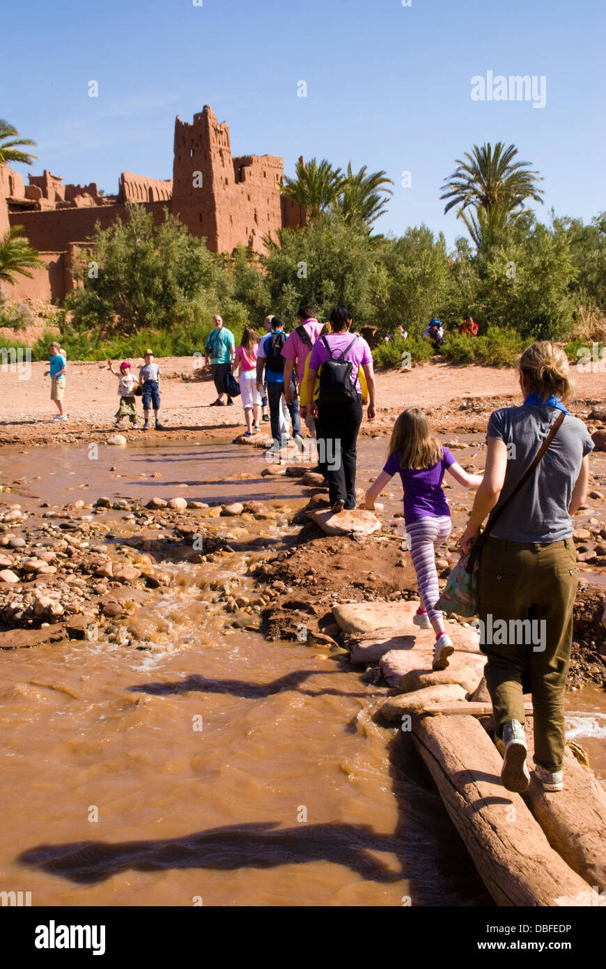 Aït Benhaddou in Berber Ath Benhadu Morocco. Tourists cross the Ounila River. Most of the town's inhabitants now live in a more modern village at the other side of the river; however, eight families still live within the ksar. Stock Photo