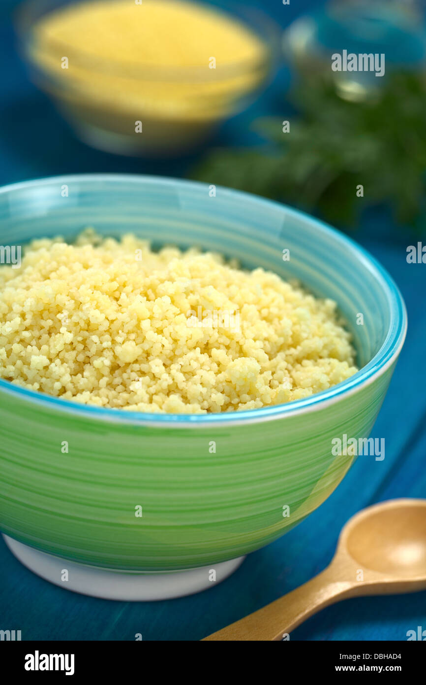 Prepared couscous in colorful bowl on blue wooden surface with raw couscous, parsley and oil in the back Stock Photo