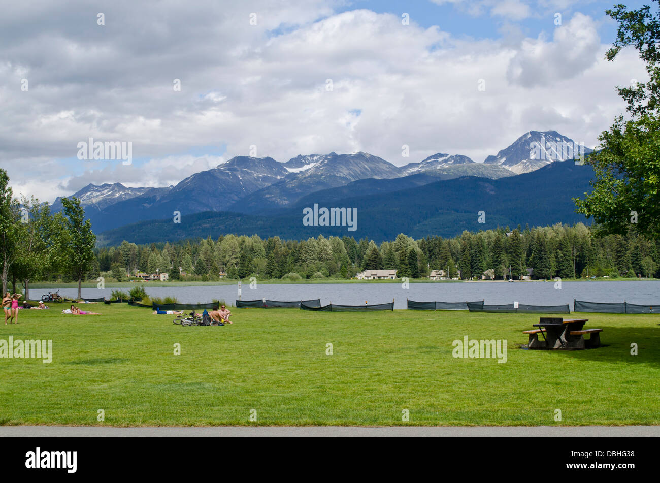 Green lawn, sunbathers and picnic tables at Rainbow Park on Alta Lake in Whistler, Canada.  Forest and mountains in the distance Stock Photo