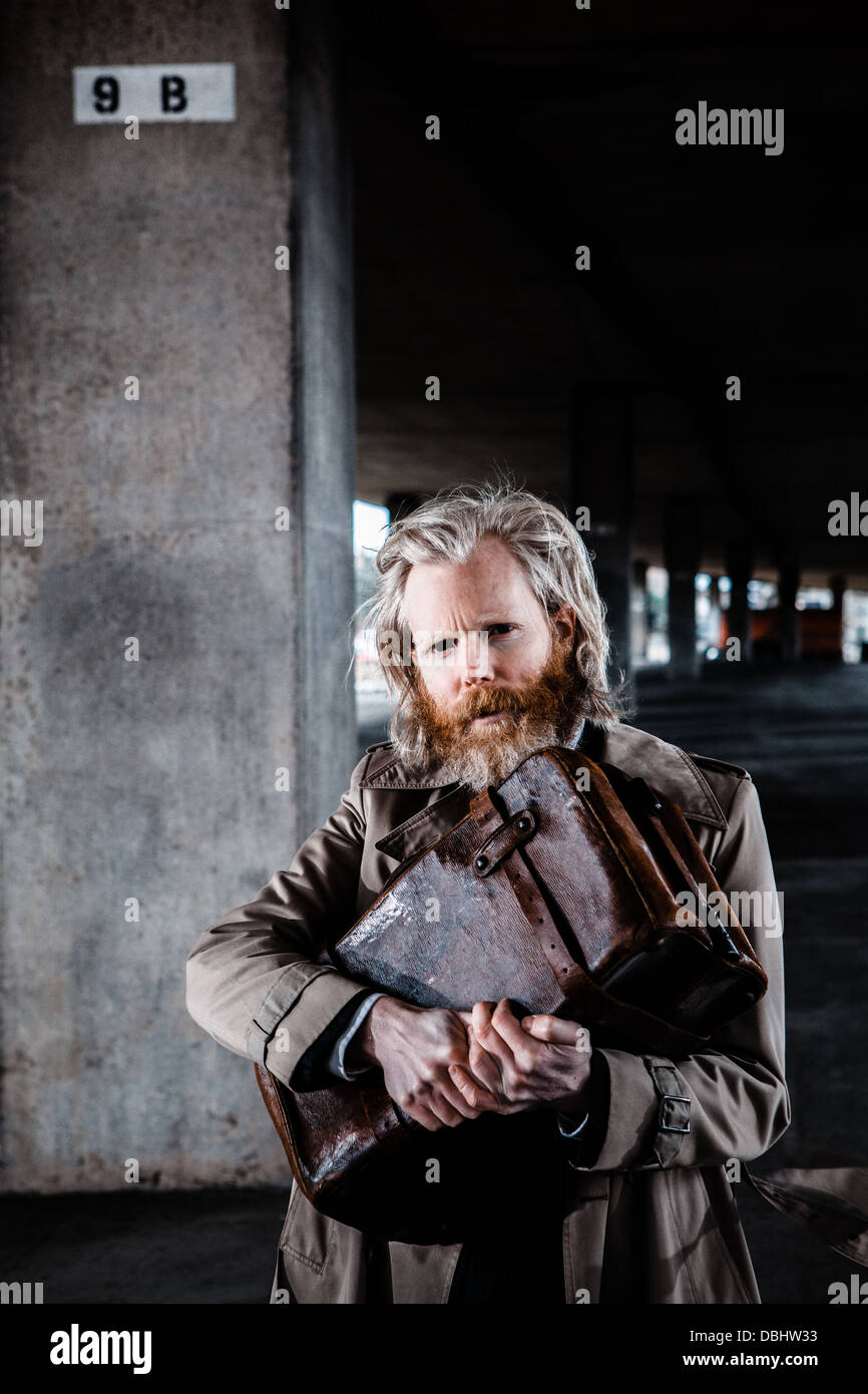 Bearded man in suit and overcoat holding an old leather briefcase in a concrete, urban setting. Stock Photo