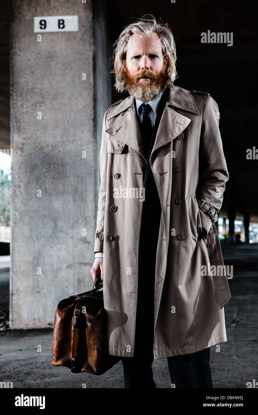 Bearded man in suit and overcoat holding an old leather briefcase standing in a concrete, urban setting. Stock Photo
