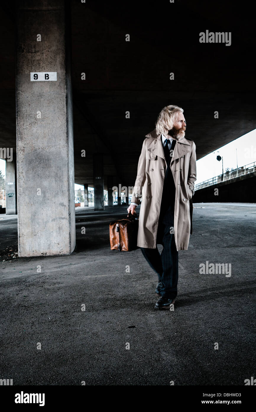 Bearded man in suit and overcoat holding an old leather briefcase walking in a concrete, urban setting. Stock Photo