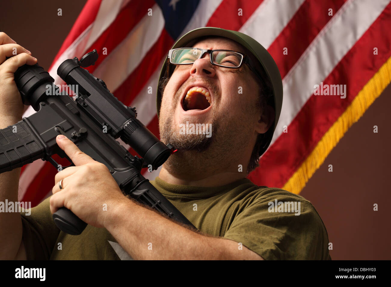 aggressive GI in t-shirt holds assault rifle with telescopic sight and screams mouth open in front of stars and stripes Stock Photo