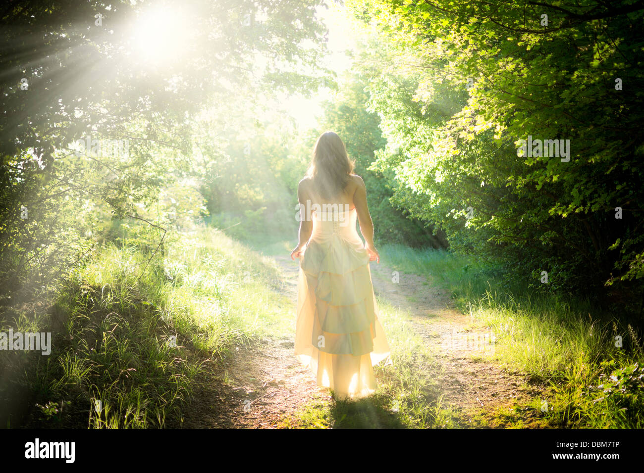 Beautiful young fairy woman walking on a path in a magical enchanted forest with dreamy beams of light shining over the road Stock Photo
