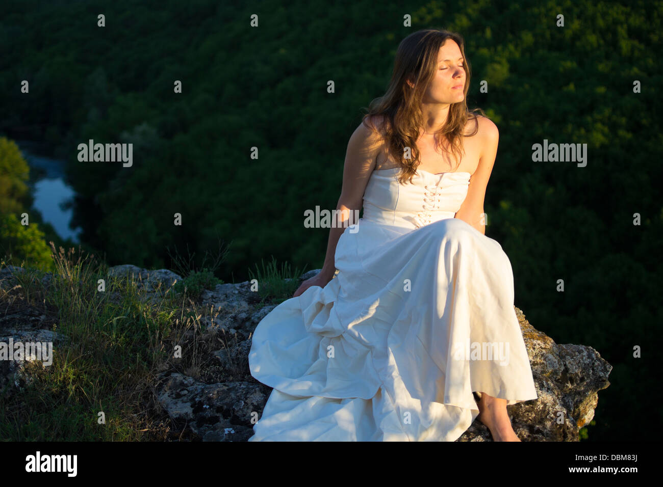 Beautiful young woman wearing elegant white dress sitting on a rock  surrounded by green forest Stock Photo