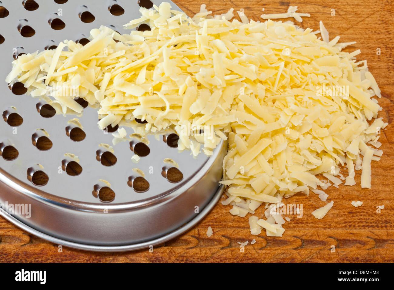 Grated Cheese - a pile of grated cheddar cheese spilling over a cheese grater, on a wooden board. Stock Photo