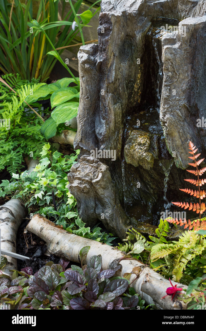 Tree stump effect resin water feature, in a damp woodland themed planting area. Stock Photo