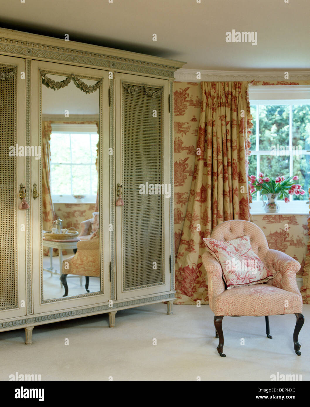 Pink armchair beside antique wardrobe with mirrored door in country bedroom with pink Toile-de-Jouy curtains and white carpet Stock Photo