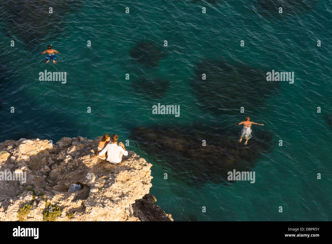 A young couple sit on a rocky outcrop overlooking swimmers in the Mediterranean sea in Nerja, southern Spain Stock Photo