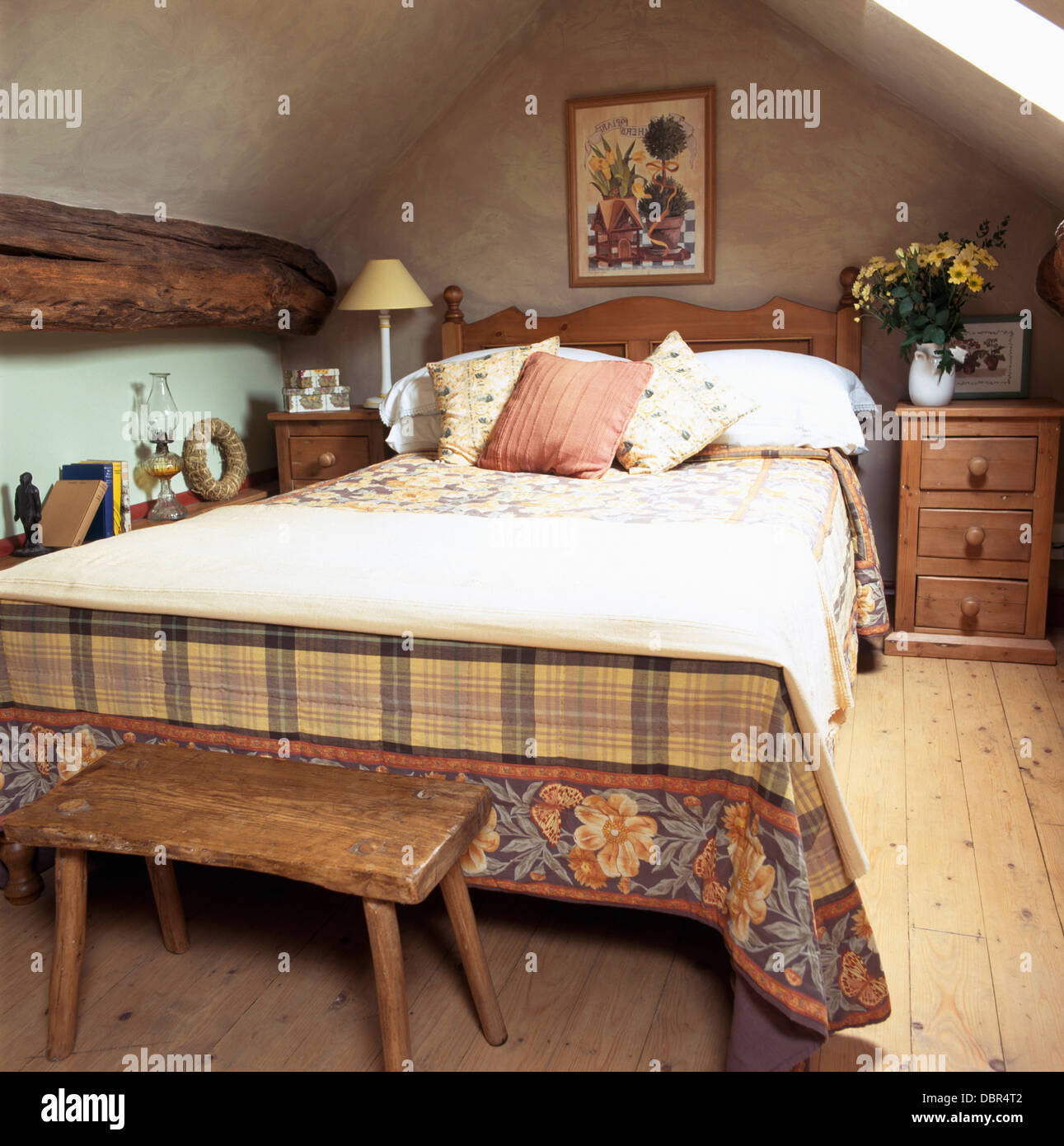 Old wooden stool below bed with cream throw and patterned bed cover in attic bedroom with wooden flooring Stock Photo