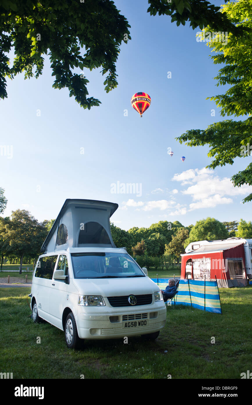 A brightly coloured hot air balloons flying above a campsite with and a VW camper van pitched up on a peaceful summer evening. Stock Photo