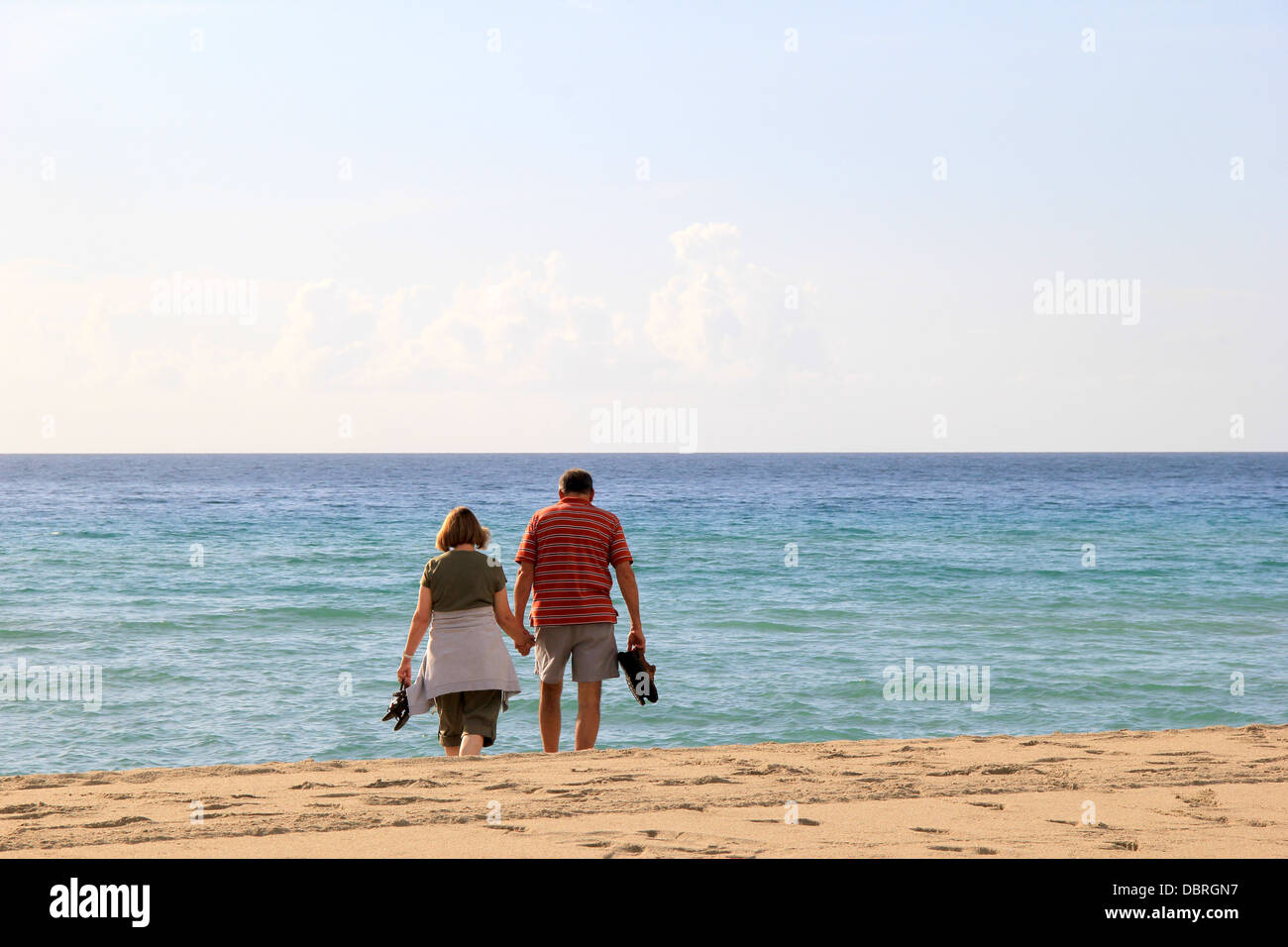 Man and woman holding shoes in one hand while other fingers are intertwined as they walk towards the water towards the shoreline Stock Photo