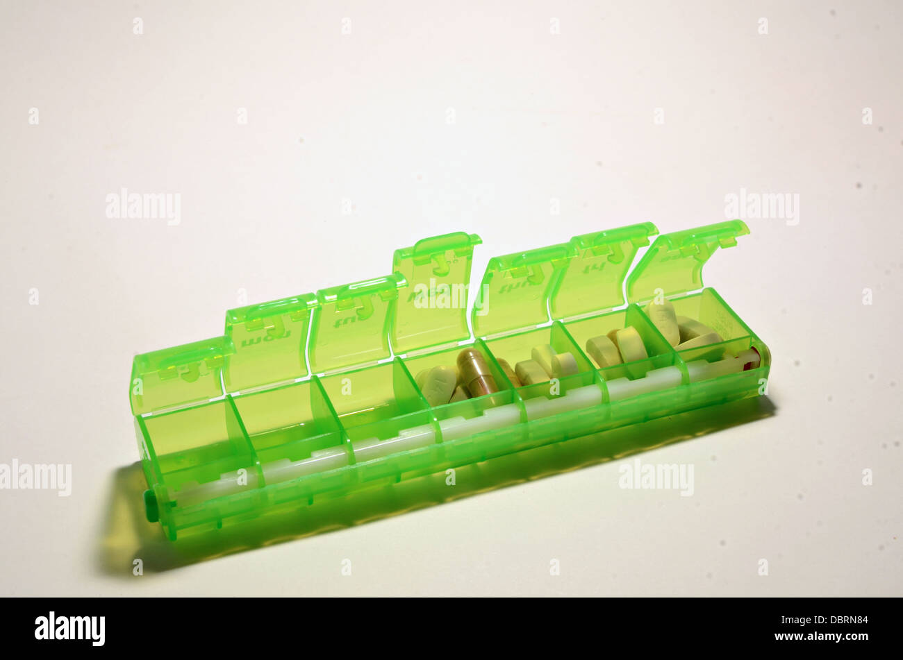 A green colored weekly pillbox sits on a white background with a assortment of pills in the box. Stock Photo