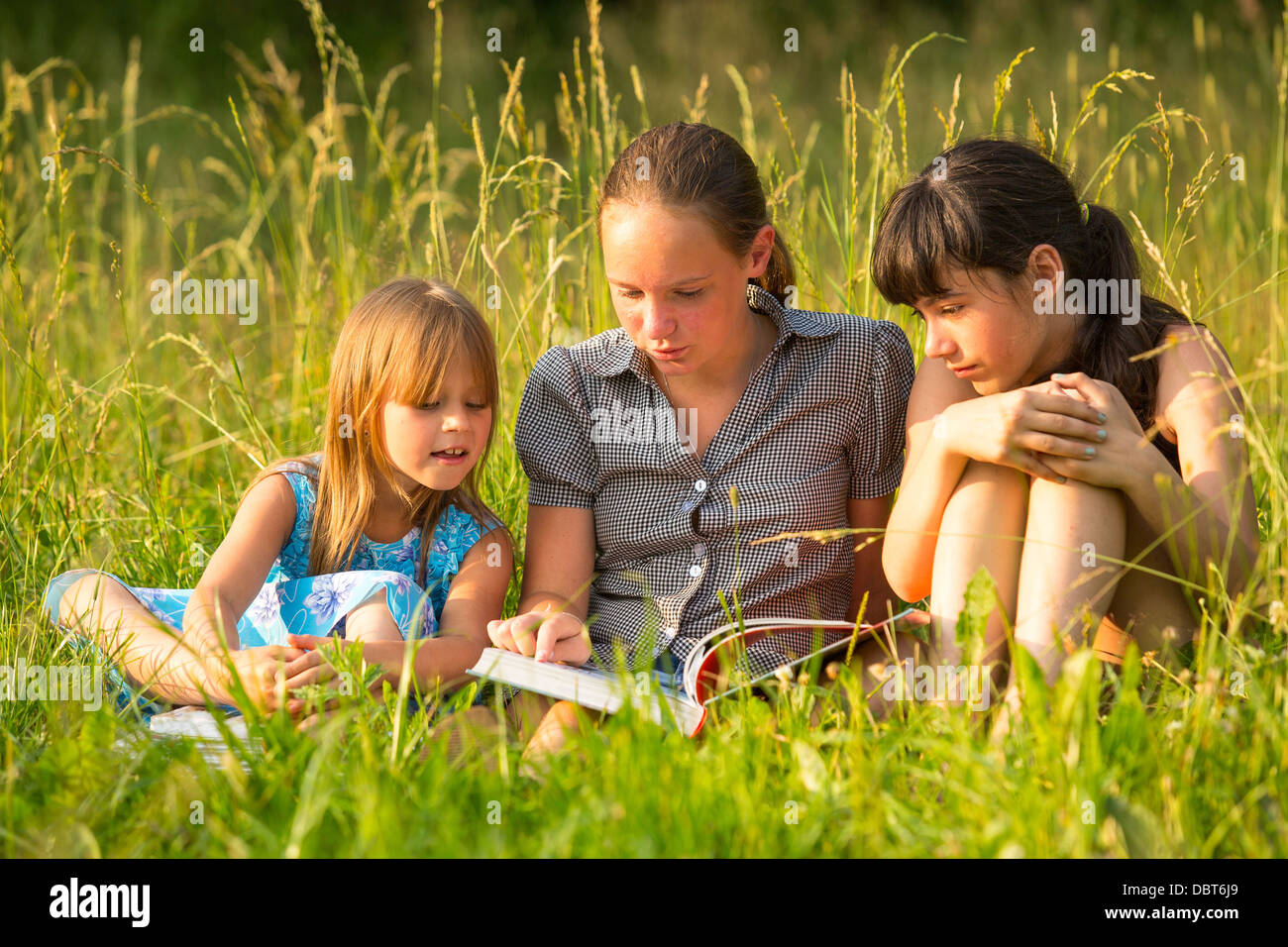 Three cute little girls reading book in natural environment together. Stock Photo