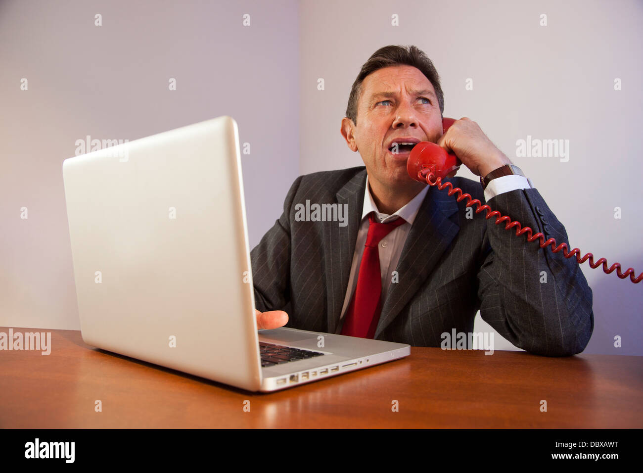 Angry man wearing a suit, shouting down a red telephone while sitting in front of a laptop on a desk. Stock Photo