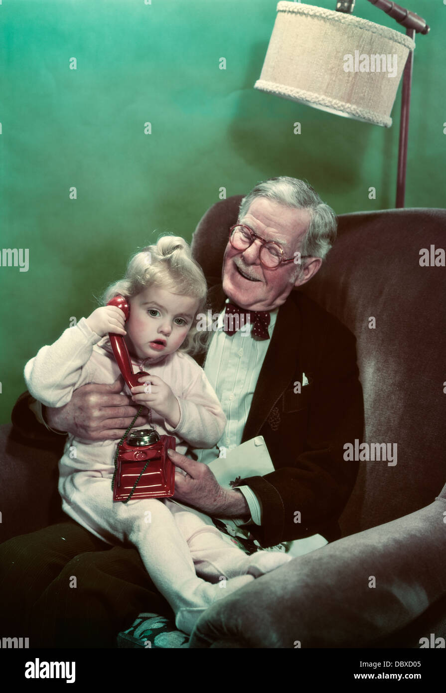 1940s 1950s GRANDDAUGHTER SITTING ON SMILING GRANDFATHER’S LAP TALKING ON TOY TELEPHONE Stock Photo