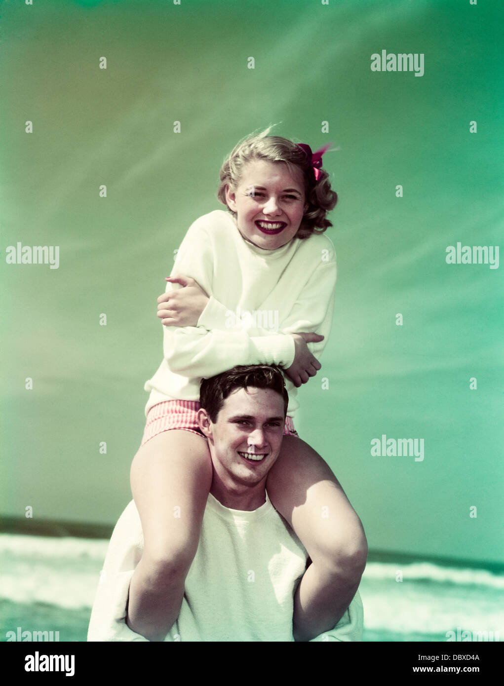 1940s 1950s SMILING TEEN COUPLE AT BEACH GIRL SITTING ON BOYS SHOULDERS LOOKING AT CAMERA Stock Photo
