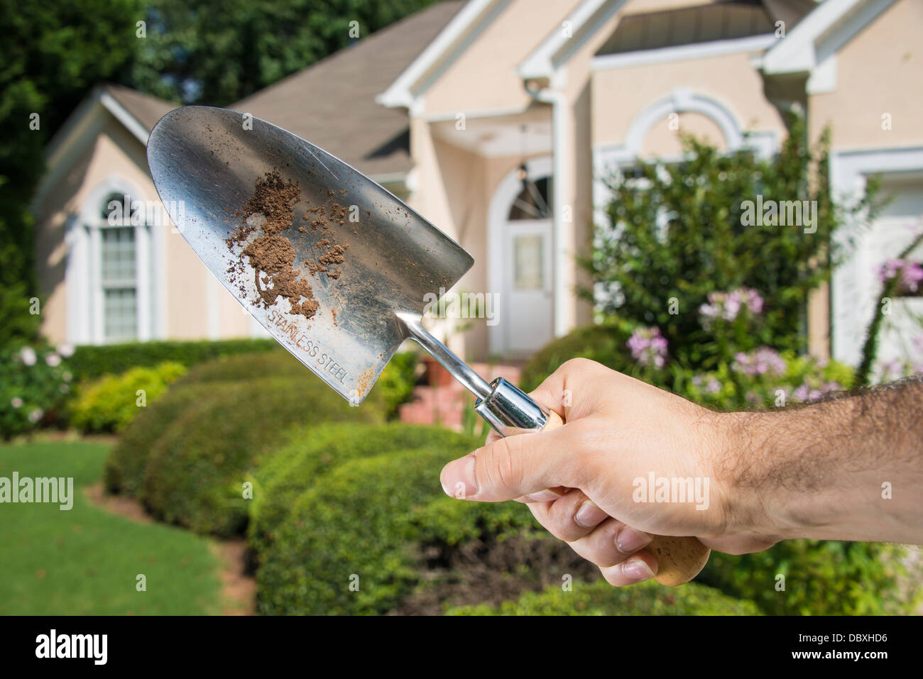Man's hand holding dirty gardening spade in front of a home's front lawn. Stock Photo