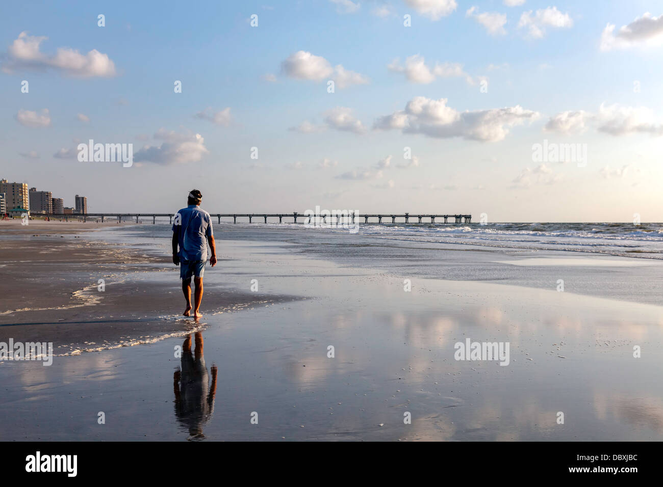 Man walking along beach at edge of the surf at dawn with fishing pier visible beyond. Stock Photo